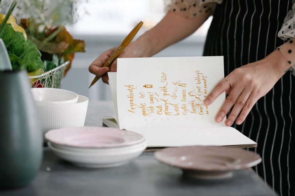 Woman writing a recipe in a notebook in a kitchen setting, surrounded by kitchenware.