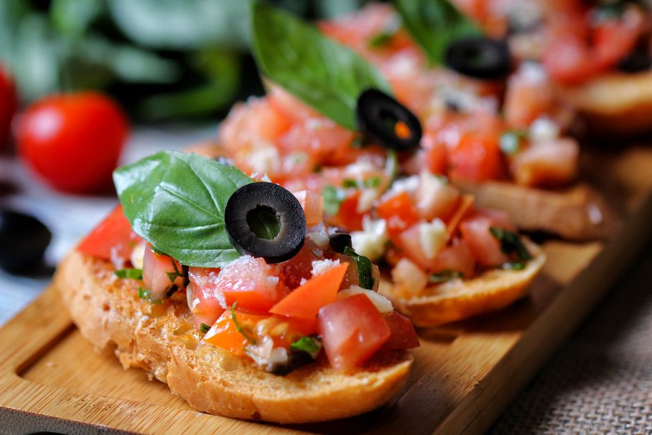 Close-up of Italian bruschetta topped with basil, tomatoes, and black olives on wooden board.